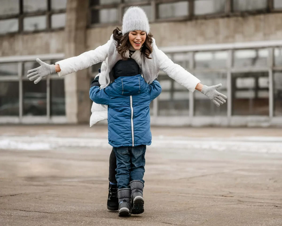 Uma mulher vestida com um casaco de frio branco e calça preta abraça uma criança com um casaco de frio azul, calça jeans e bota de inverno infantil em um ambiente urbano