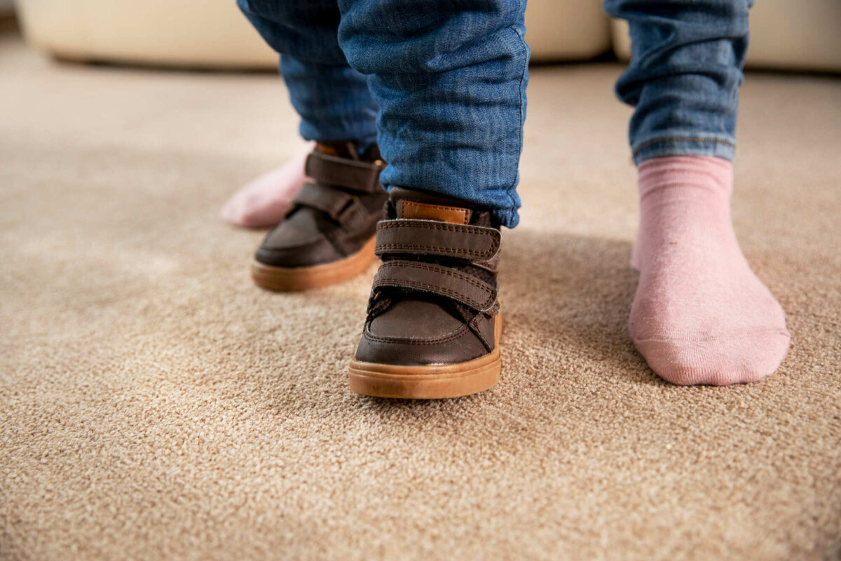 Pés de bebê calçados com sapatos infantis em um carpete marrom e pés do pai atrás