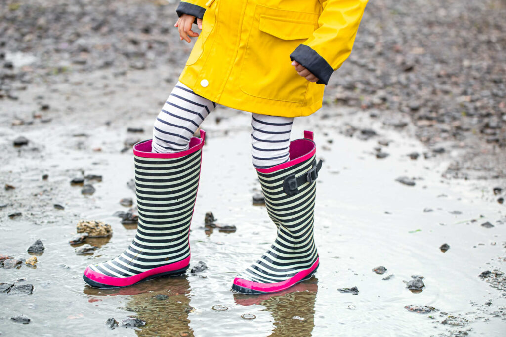 Uma criança vestida com um casaco de chuva amarelo, calça listrada em preto e branco e galochas listradas em preto e branco com detalhe rosa em pé em uma poça d'água