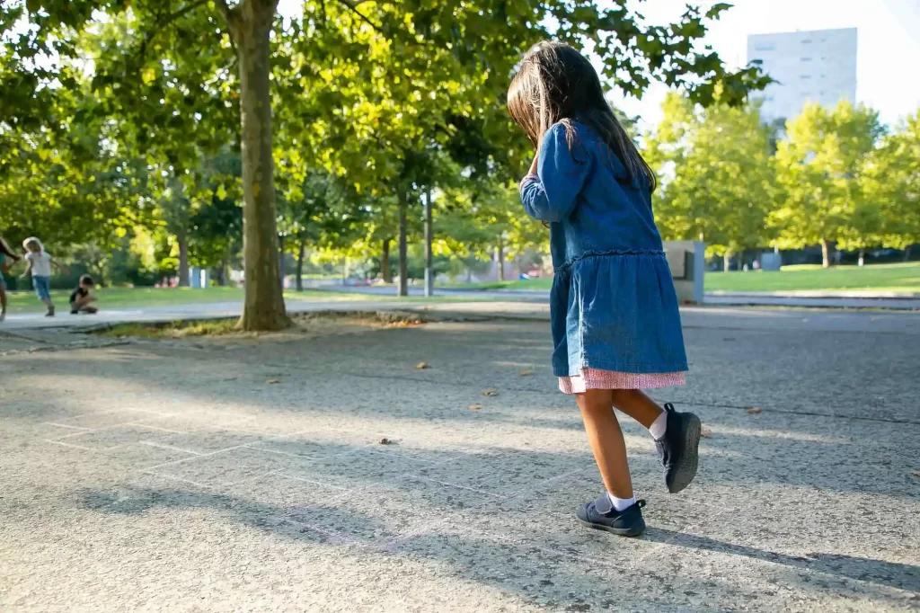 Menina brincando de amarelinha desenhada no chão com giz em um parque. Ela usa um vestido jeans e sapatos pretos, enquanto outras crianças brincam ao fundo entre as árvores e áreas verdes. O sol brilha através das folhas, iluminando a cena.