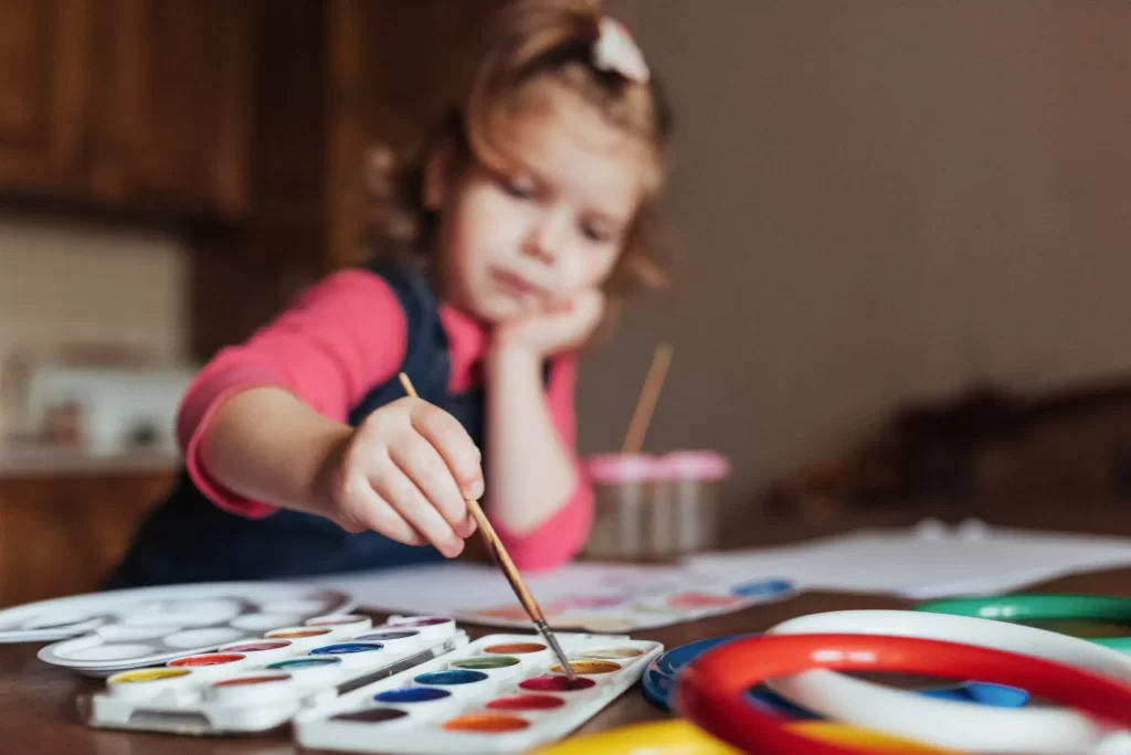 Uma menina pequena está concentrada em uma atividade de pintura. Ela está segurando um pincel, mergulhando-o em uma paleta de aquarela colorida. Ao fundo, a menina está desfocada, vestindo uma camiseta rosa e avental. A mesa está coberta com materiais de arte, incluindo tintas e papéis.