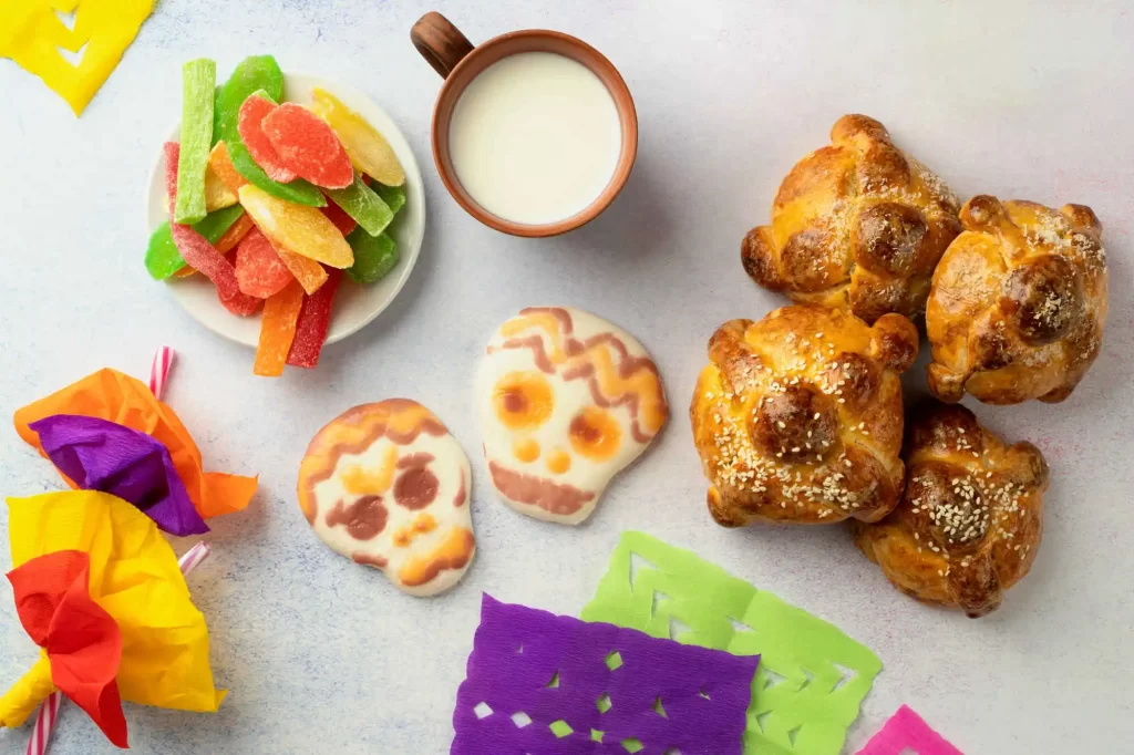 Uma mesa com um café da manhã. Há pães doces conhecidos como 'pan de muerto', biscoitos em forma de caveira, uma tigela de leite, frutas cristalizadas coloridas e decorações de papel recortado em cores vivas.