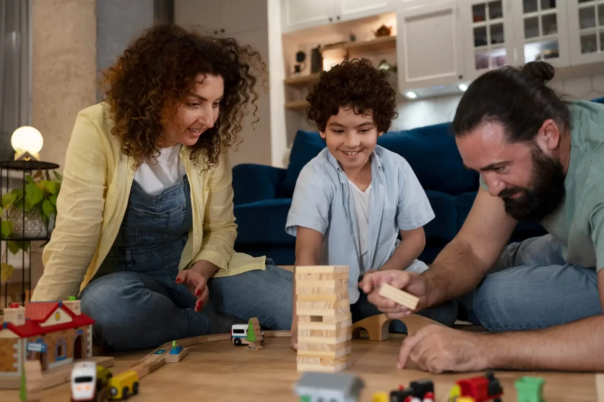 Um menino brincando com os pais no chão de uma sala de estar. Eles estão sorrindo e se divertindo enquanto constroem uma torre de blocos de madeira e brincam com um conjunto de trens e carros de brinquedo. Ao fundo, há uma sala bem decorada com um sofá azul e prateleiras.