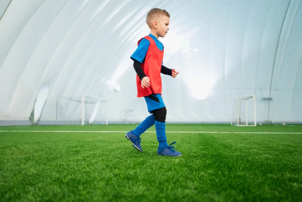 Imagem de uma criança usando uma chuteira infantil de campo em um campo de futebol fechado usando um uniforme azul com preto e colete laranja