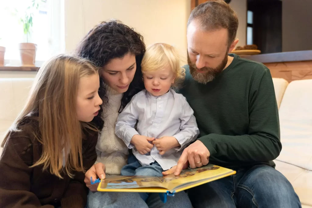 Uma família sentada em um sofá, lendo um livro infantil. O pai, com barba e usando uma camiseta verde, está segurando o livro e apontando algo para o filho mais novo, que está no colo dele, vestindo uma camisa azul clara. A mãe, com cabelos cacheados escuros, está sentada ao lado, olhando para o livro, enquanto a filha mais velha, de cabelos longos e loiros, está ao lado dela, também atenta à leitura. O ambiente é aconchegante e bem iluminado.