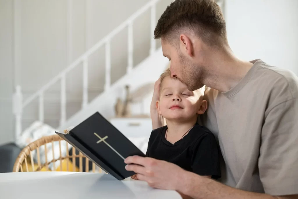 Um pai está sentado com seu filho pequeno, segurando uma bíblia. O pai está beijando carinhosamente a cabeça do filho, que está com os olhos fechados e uma expressão de contentamento. Eles estão em um ambiente aconchegante, com uma escada ao fundo. 