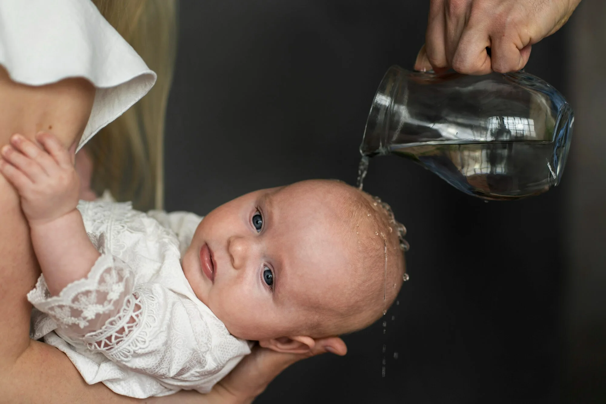 Um bebê vestido com uma roupa branca rendada está sendo segurado por um adulto, enquanto outra pessoa derrama água sobre a cabeça do bebê usando uma jarra de vidro, realizando o batismo. A expressão do bebê é calma e atenta, e o fundo da imagem é escuro, destacando o bebê e a jarra.