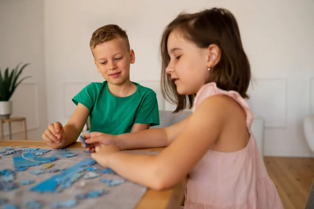 Duas crianças montando um quebra-cabeça. A menina, vestindo um vestido rosa, está focada em encaixar as peças, enquanto o menino, vestindo uma camiseta verde, observa e ajuda. Eles estão sentados em uma mesa em um ambiente doméstico bem iluminado.