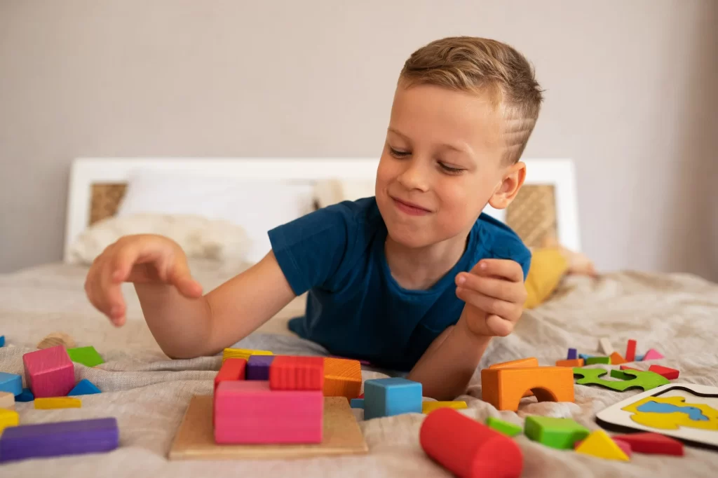 Um menino loiro deitado na cama, brincando com blocos de construção coloridos. Ele está usando uma camiseta azul e sorrindo levemente enquanto organiza os blocos em uma base de madeira. Ao redor dele, há vários blocos de diferentes formas e cores, além de um quebra-cabeça parcialmente montado.