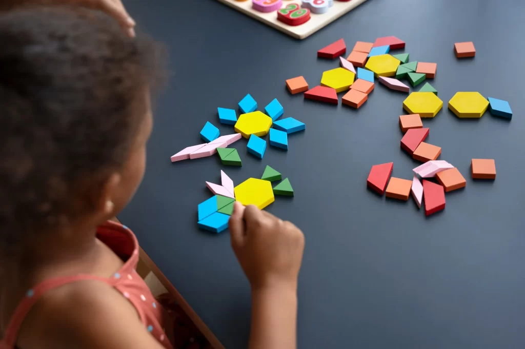 Uma criança brincando com blocos coloridos de formas geométricas em uma mesa preta. A criança está criando desenhos com os blocos, formando flores e outras formas criativas. Ao fundo, há mais brinquedos educativos sobre a mesa.