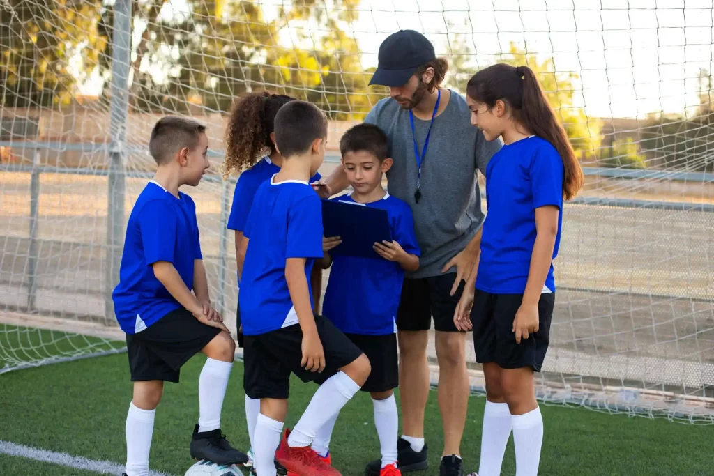 Um grupo de cinco crianças vestidas com uniformes de futebol azul e branco está reunido ao redor de um treinador em um campo de futebol. O treinador, usando boné e camiseta cinza, está segurando uma prancheta e conversando com as crianças. Ao fundo, uma rede de gol e algumas árvores são visíveis.