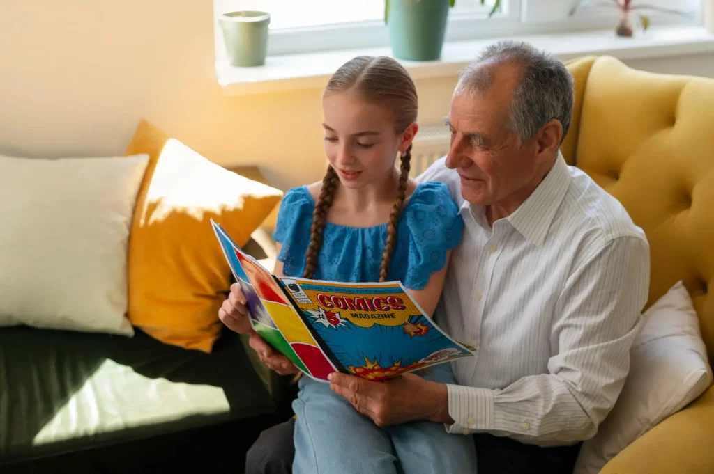 Um avô e sua neta estão sentados juntos em um sofá amarelo, lendo uma revista em quadrinhos. A menina, com tranças e vestindo uma blusa azul, está sorrindo enquanto segura a revista. O avô, vestindo uma camisa branca, olha para a revista com interesse. Ao fundo, há uma janela com luz natural iluminando a cena e almofadas coloridas no sofá.