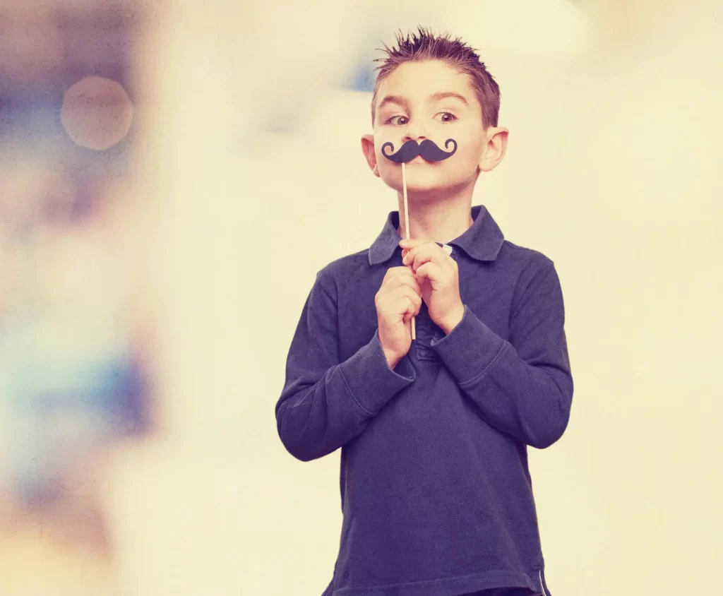 Um menino pequeno está segurando um bigode de papel preto preso a um palito em frente ao rosto, como se estivesse fingindo ter um bigode. Ele veste uma camisa polo azul e olha para o lado com uma expressão divertida. O fundo é claro e desfocado, destacando o menino na imagem.