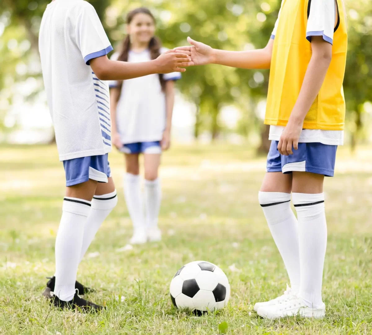 Três crianças em uniformes de futebol estão ao ar livre, com duas delas apertando as mãos e uma bola de futebol no gramado à frente delas. A terceira criança, em segundo plano, observa a interação. As crianças estão em um campo gramado com árvores ao fundo, indicando um ambiente de jogo ou treino de futebol