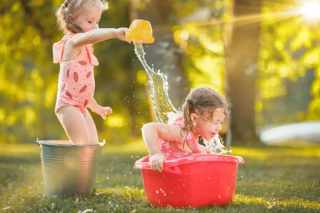 Duas crianças brincando com água em um jardim. Uma menina está de pé dentro de um balde de metal, vestindo um maiô rosa, e derramando água de um pequeno balde amarelo sobre a cabeça da outra menina. A outra menina, também de maiô rosa, está inclinada dentro de uma bacia vermelha, rindo enquanto a água cai sobre ela. O fundo mostra um jardim com árvores e luz do sol brilhando através das folhas.