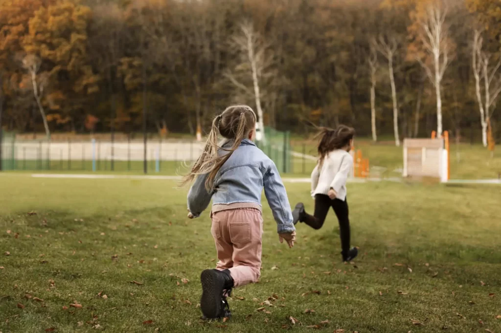 Duas crianças correndo em um campo de grama verde em um parque. A criança mais próxima à câmera usa jaqueta jeans e calça rosa, enquanto a outra criança usa um casaco claro e calça preta. Ao fundo, há árvores com folhas outonais e equipamentos de playground.