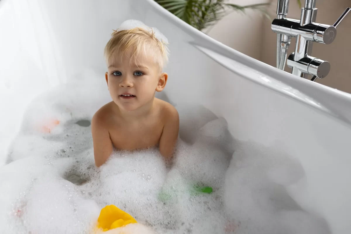 Um menino pequeno está brincando em uma banheira cheia de espuma. Ele está sentado na banheira, cercado por bolhas de sabão e alguns brinquedos de banho coloridos. A expressão do menino é atenta e curiosa. O ambiente é iluminado e moderno, com uma torneira cromada visível ao lado da banheira.