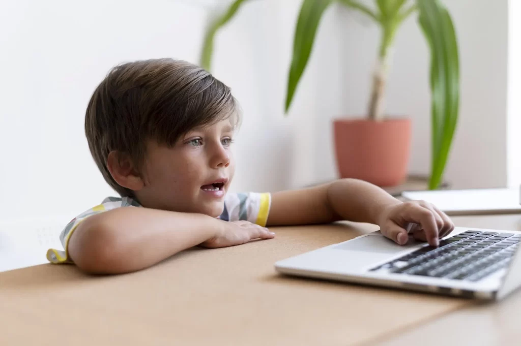 Um menino pequeno está sentado em frente a um laptop, com o braço apoiado na mesa e a mão no teclado. Ele olha atentamente para a tela enquanto usa o computador. Ao fundo, há uma planta em um vaso laranja em cima da mesa. A luz natural ilumina a cena, destacando o foco do menino.