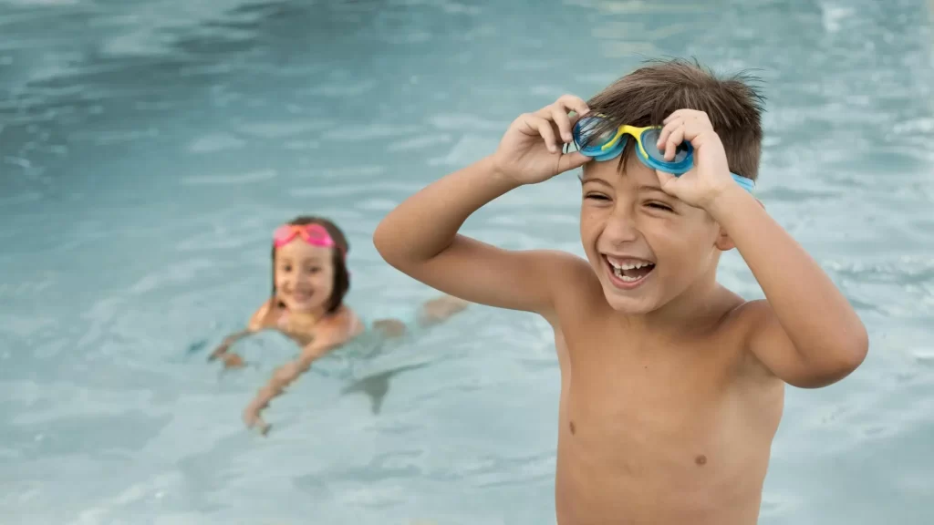 Duas crianças brincando em uma piscina. Um menino está em primeiro plano, sorrindo amplamente enquanto ajeita seus óculos de natação azuis na cabeça. Ao fundo, uma menina está na água, também sorrindo e usando óculos de natação rosa.