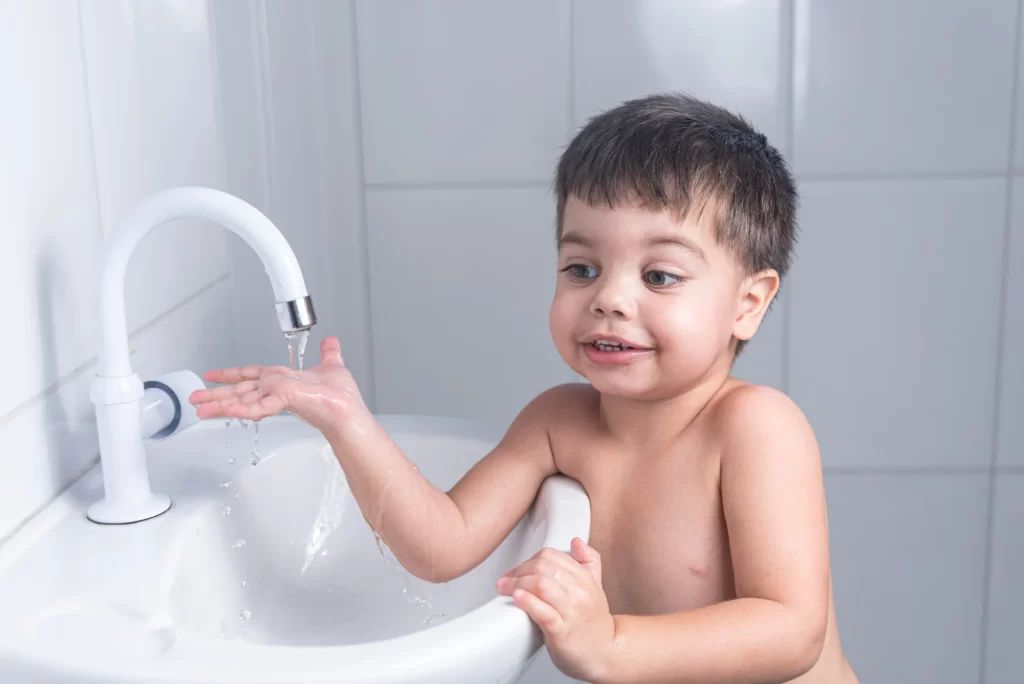 Menino pequeno, sem camisa, lavando as mãos em uma pia de banheiro branco, olhando curioso para a água que sai da torneira.