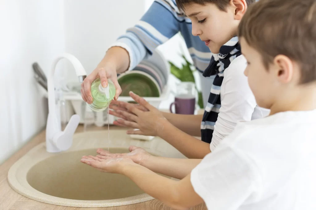 Dois meninos lavando as mãos em uma pia, com a ajuda de um adulto que está derramando sabonete líquido nas mãos deles. Os meninos estão concentrados, um olhando para suas mãos e o outro para o sabão.