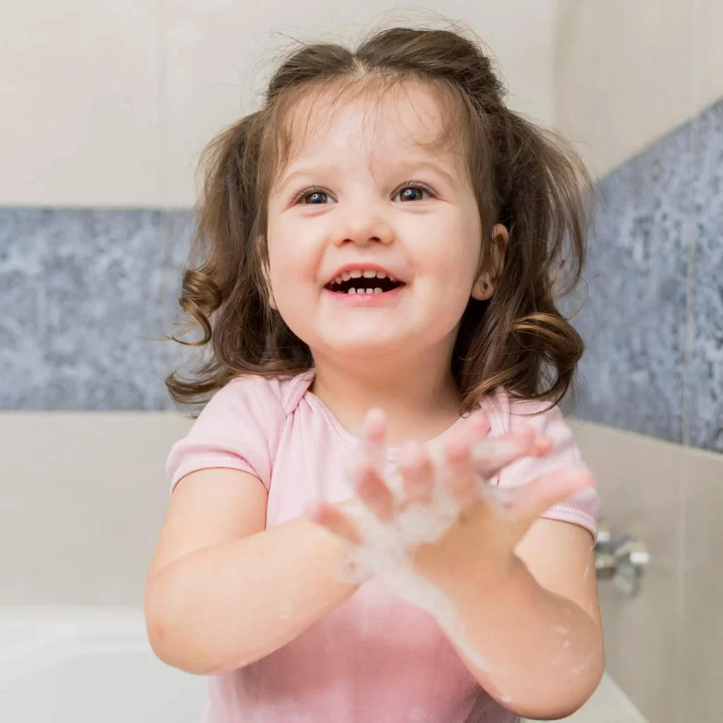 Uma menina sorridente com cabelo castanho encaracolado está lavando as mãos em um banheiro. Ela usa uma camiseta rosa e suas mãos estão cobertas de espuma de sabão.