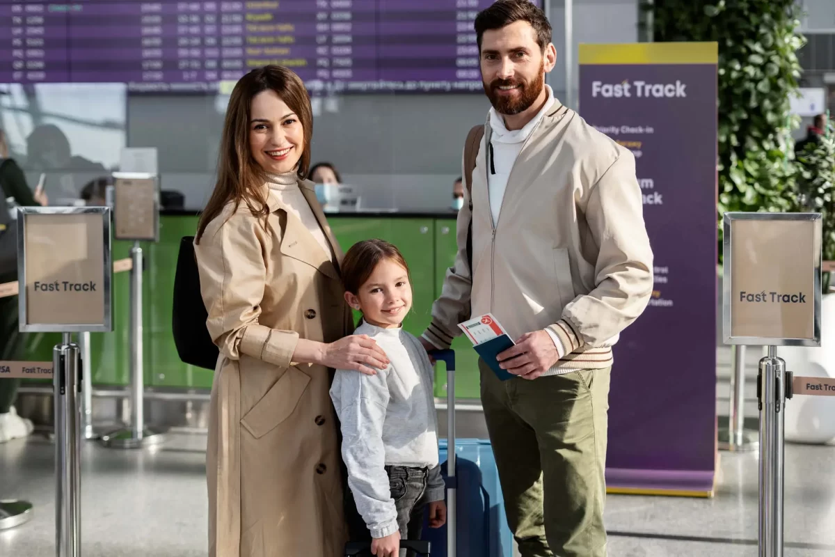 Uma família de três pessoas, composta por um homem, uma mulher e uma criança, está no aeroporto. Eles estão na área de check-in rápido, segurando passaportes e cartões de embarque. Todos estão sorrindo e parecem prontos para viajar. A mulher está vestindo um casaco bege, a criança um suéter cinza e o homem um casaco claro. Atrás deles, há sinais que dizem "Fast Track" e uma tela de informações de voos.