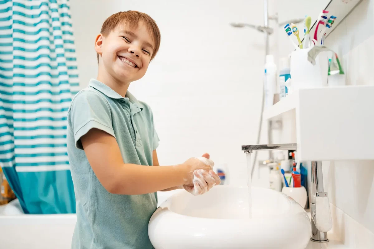 Menino sorridente lavando as mãos com sabão em uma pia de banheiro, com cortina de chuveiro listrada azul e branca ao fundo.