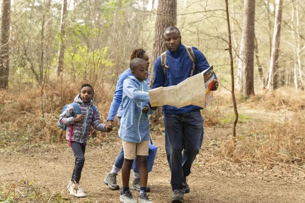 Família fazendo trilha em uma floresta, com o pai segurando um mapa e guiando o caminho, enquanto a mãe e duas crianças caminham ao lado, todos vestidos com roupas apropriadas para caminhada.