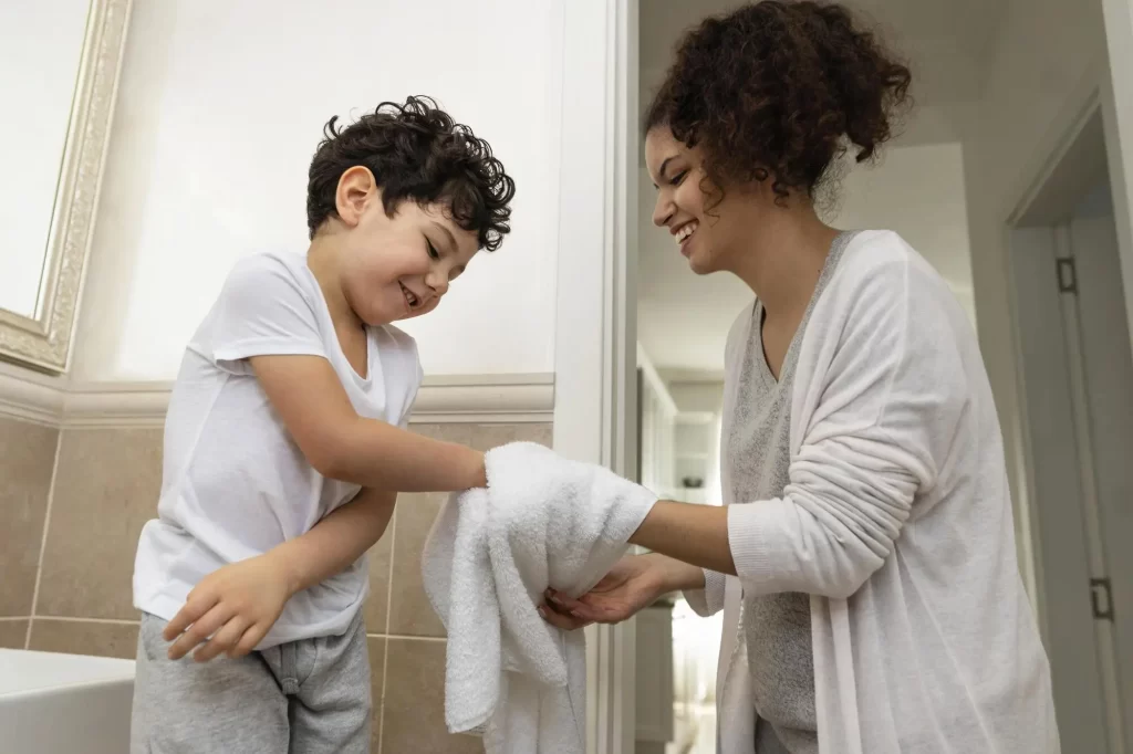 Uma criança sorridente está secando as mãos com uma toalha branca, enquanto uma mulher ao lado dela ajuda no processo. Ambos estão sorrindo, transmitindo um sentimento de alegria e cuidado. A cena ocorre em um banheiro bem iluminado, com azulejos bege e uma pia visível ao fundo.